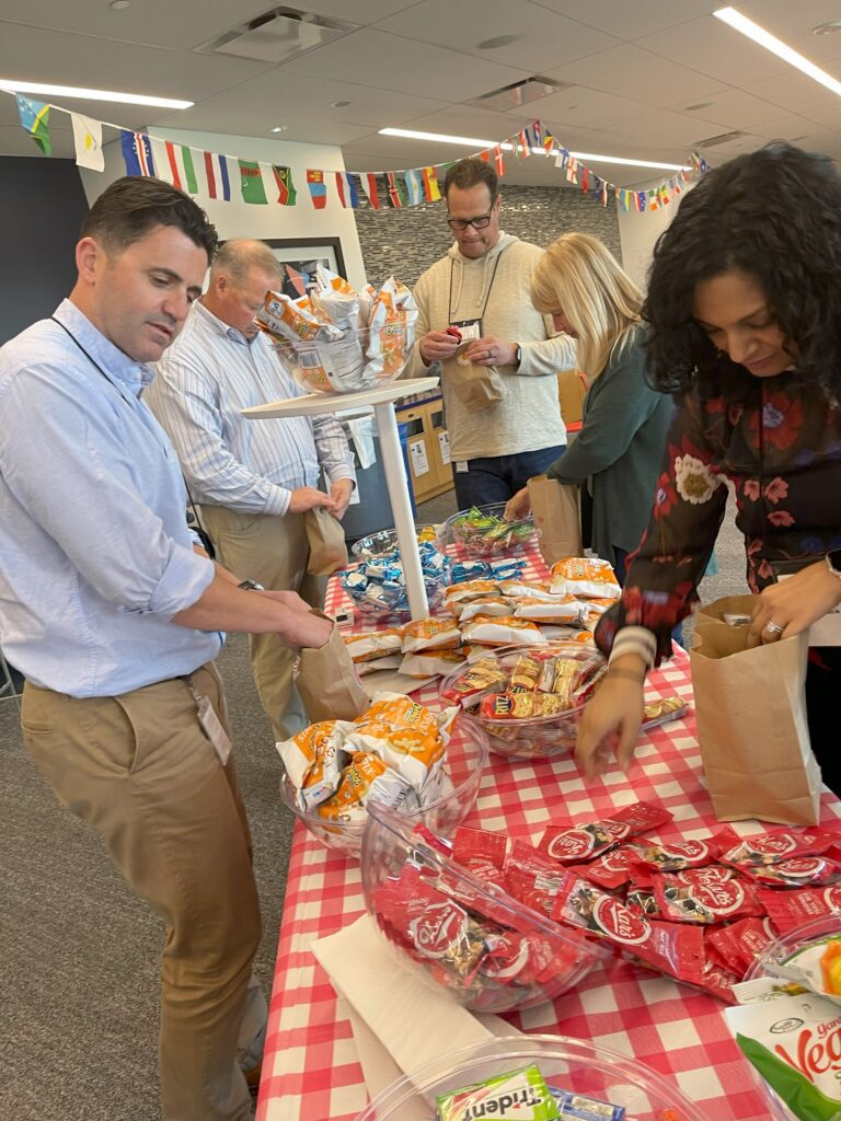 Group of people filling brown paper bags with snacks during The Great Snack Pack activity at a corporate team building event, with a table covered in a red checkered cloth and bowls of snacks.