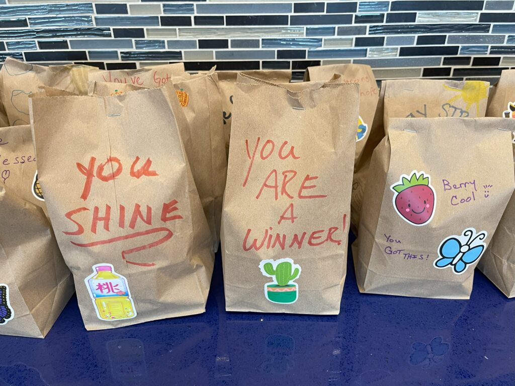 Decorated brown paper bags with motivational messages like 'You Shine' and 'You Are a Winner!' alongside stickers, prepared for The Great Snack Pack team building Donation Station.