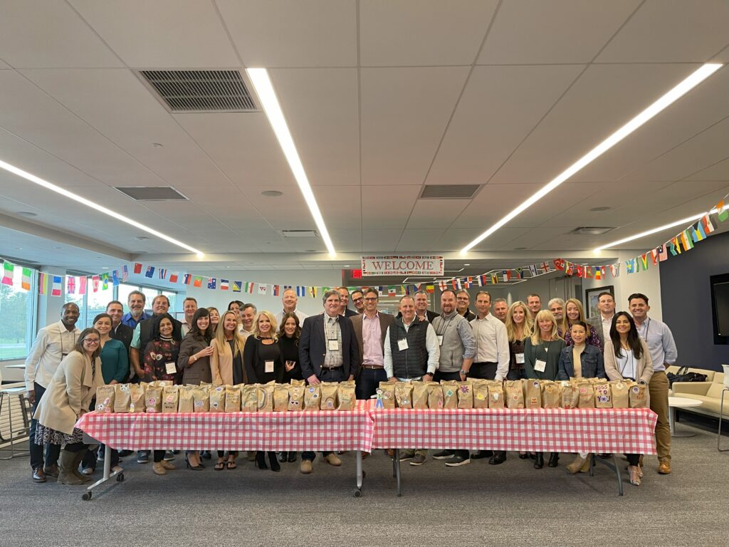 Large group of people standing behind a table filled with decorated snack bags during The Great Snack Pack activity at a corporate charitable team building event.