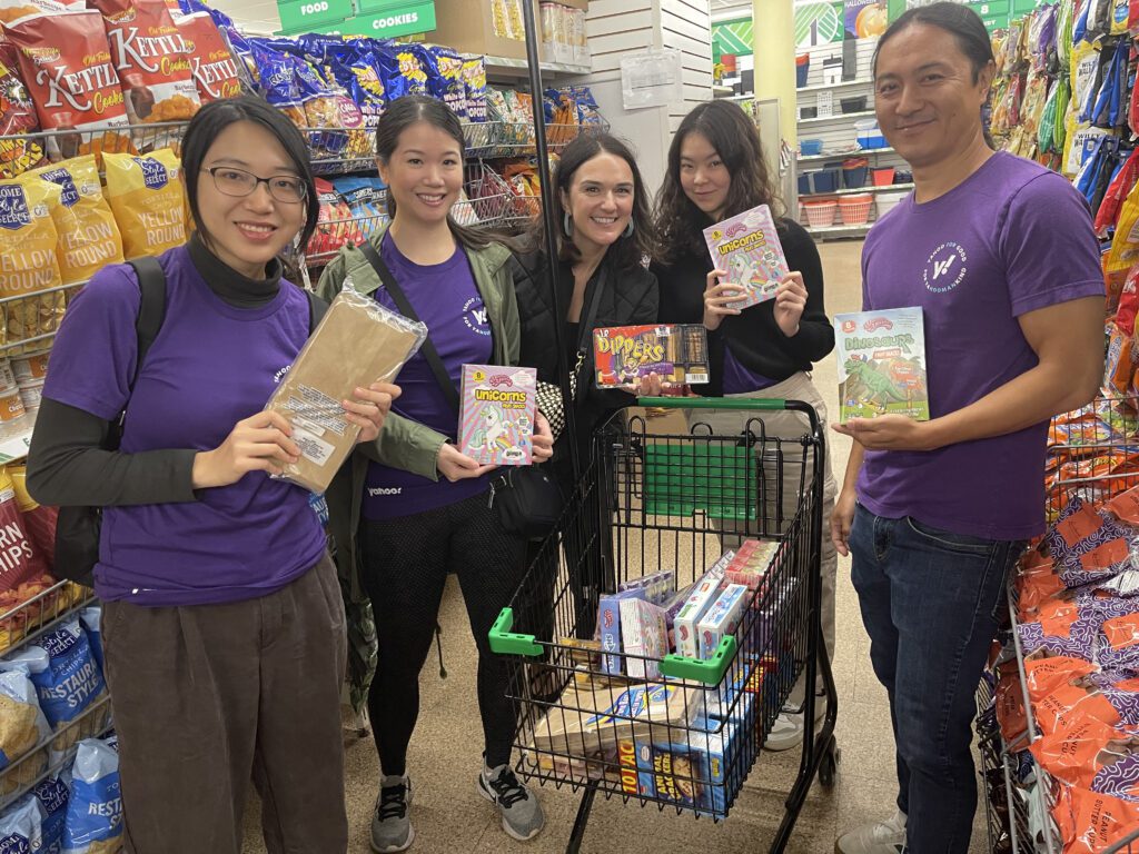 A group of five volunteers stands together in a grocery store aisle, smiling and holding up various food items. They have a shopping cart filled with packaged goods for a donation or charity event. They are participating in a Do Good Bus team building event where they are collecting items to support a community cause. The atmosphere appears cheerful and collaborative.