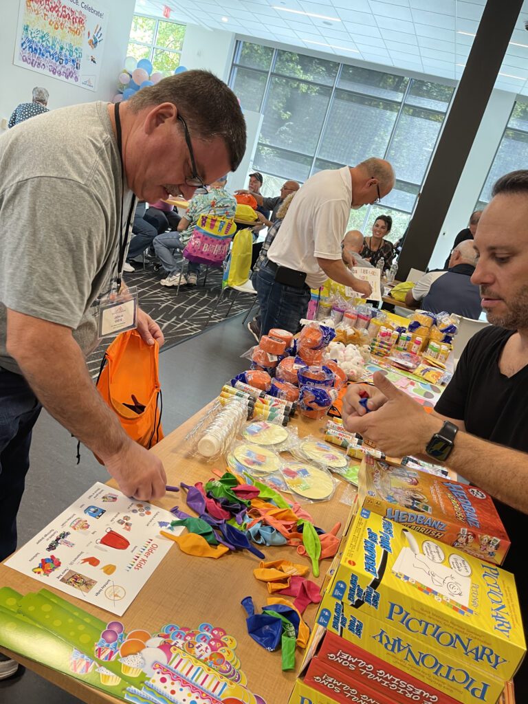 Two men are selecting colorful balloons, party supplies, and games from a table filled with items like 