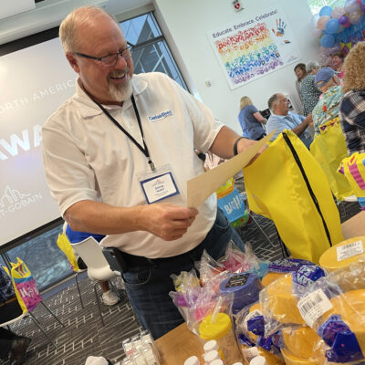 A man wearing glasses and a white shirt is smiling as he reads a card. In front of him is a table filled with colorful party supplies, decorations, and gift items for a charitable birthday-themed team building event.