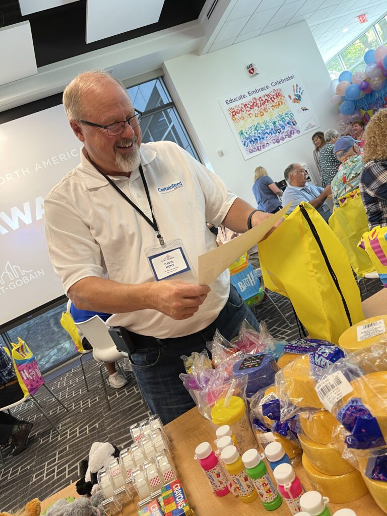 A man wearing glasses and a white shirt is smiling as he reads a card. In front of him is a table filled with colorful party supplies, decorations, and gift items for a charitable birthday-themed team building event.