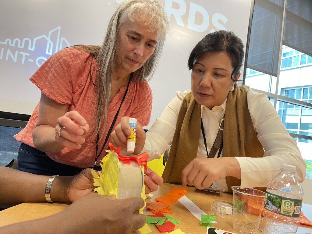 Two women are working together, focused on crafting a colorful project using glue and paper. They are engaged in a team building activity at a table with various supplies to create a piñata as part of a birthday kit donation.