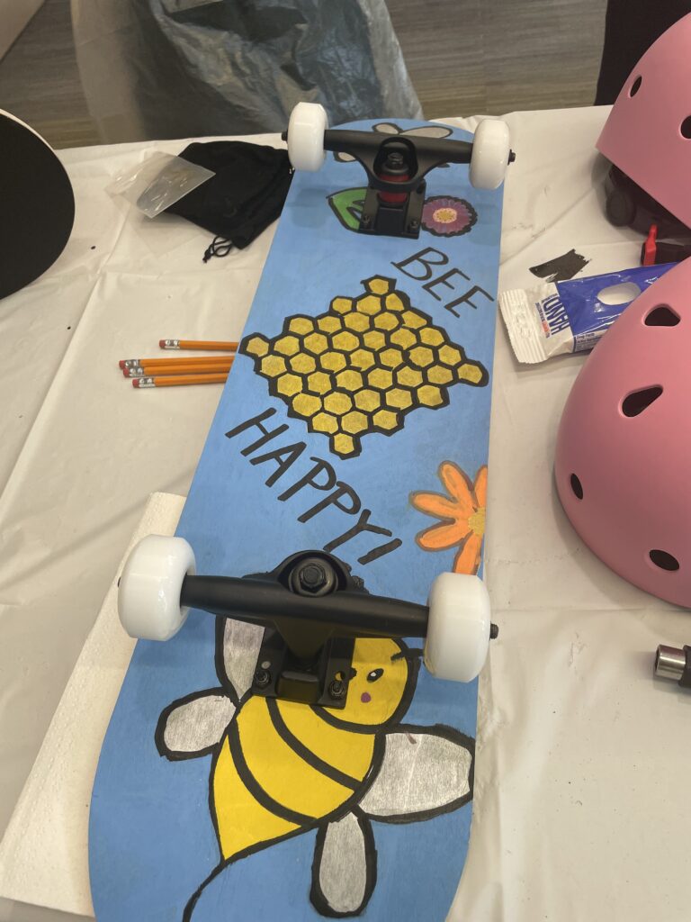 A custom-decorated skateboard featuring a cheerful design with a honeycomb, a bee, and the phrase 'BEE HAPPY!' The board has white wheels, and next to it on the table are some pencils, a pink helmet, and various crafting supplies. The skateboard is part of a creative team building event where participants assemble and decorate skateboards for a charitable cause.