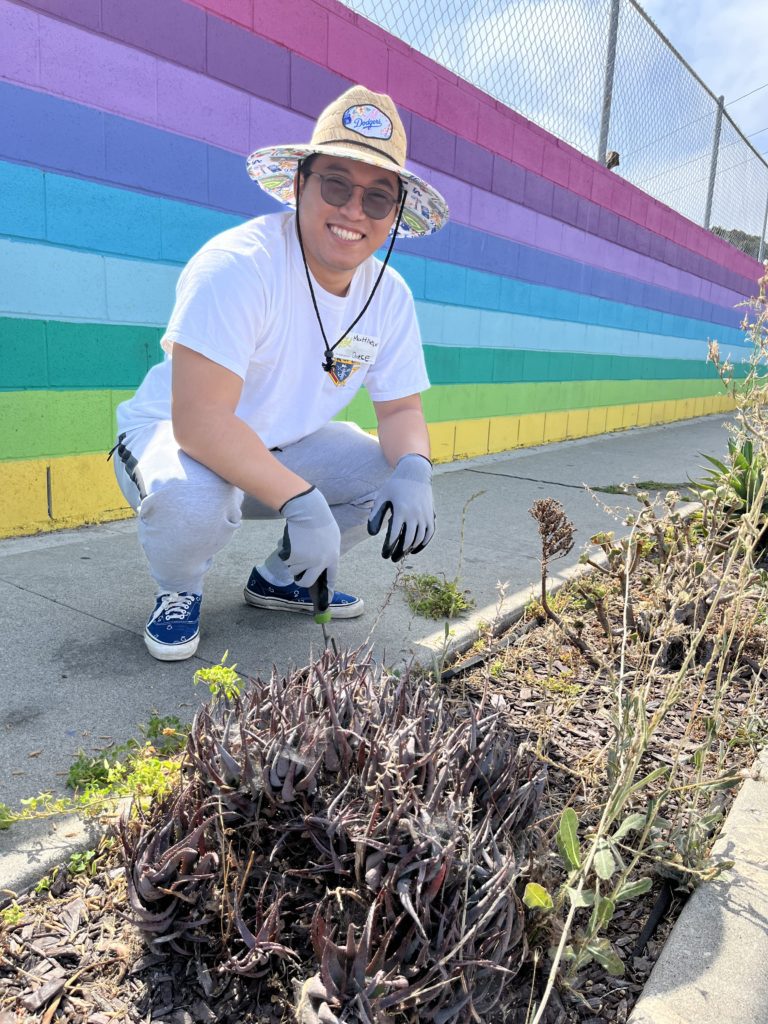 A smiling man crouches down on the sidewalk, gardening in a small flowerbed. He wears a large, colorful sun hat, sunglasses, and gardening gloves while dressed in a white T-shirt and gray sweatpants. Behind him is a vibrant rainbow-colored mural painted on a wall, creating a colorful backdrop. The man is engaged in charitable team building activity.