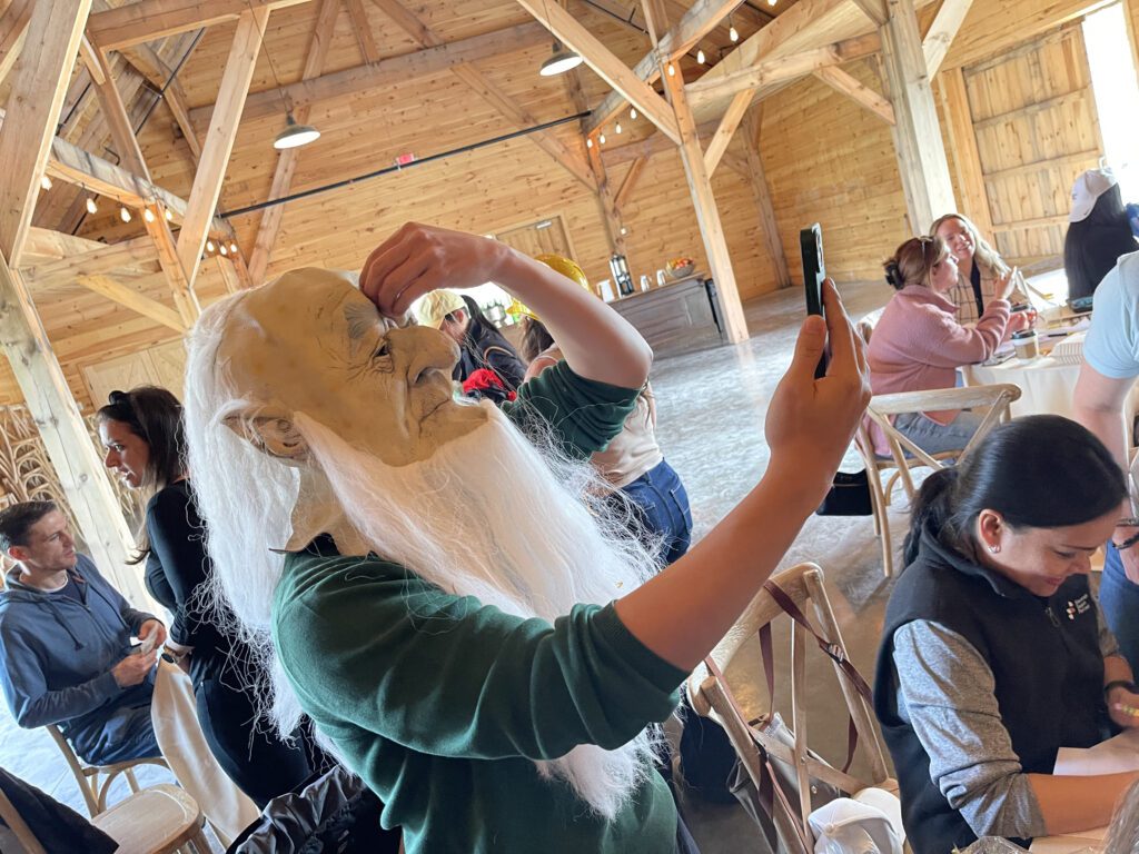 Person wearing a long white-bearded mask taking a selfie with a smartphone, preparing for the Corporate Film Festival challenge.