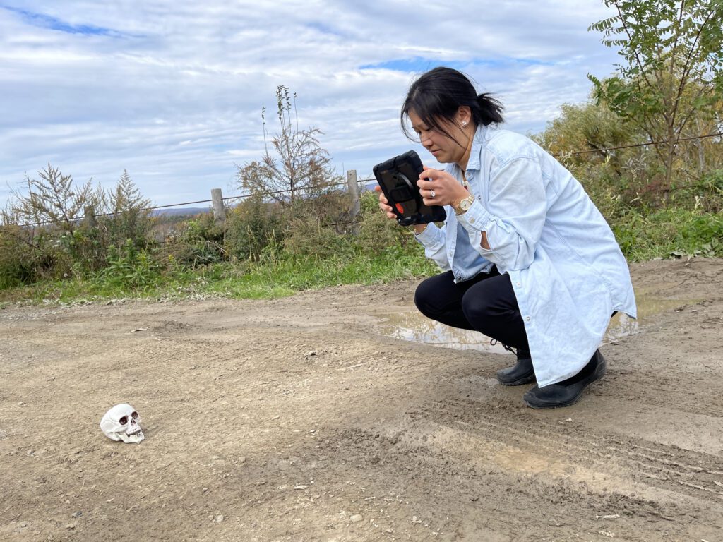 A woman crouches down on a path, using a tablet to photograph a skull placed on the ground, simulating a crime scene investigation as part of a team building activity focused on making a movie.