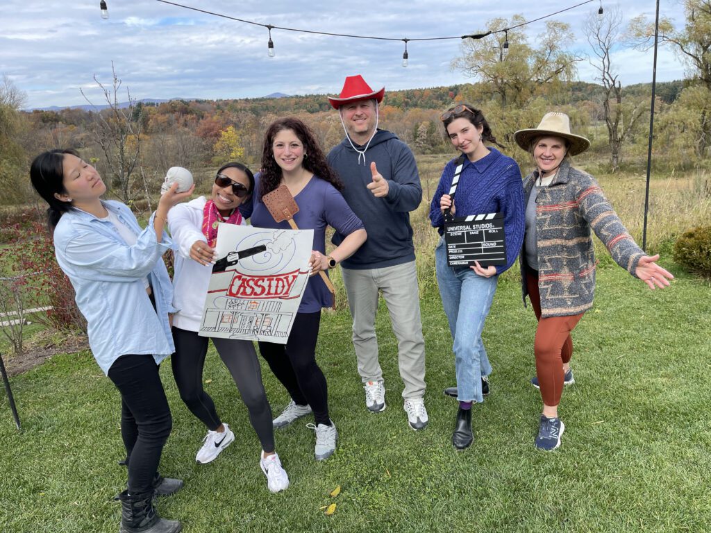 A group of six people pose outdoors, holding various props like a film clapperboard, a poster, and a red cowboy hat, participating in a creative team building activity themed around film production and storytelling.