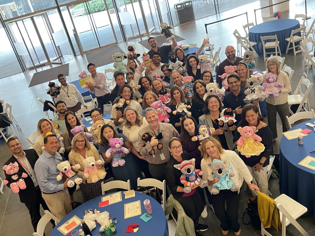 A large group of participants proudly holding their completed teddy bears at the end of the Team Teddy Rescue Bear event. The participants, arranged in a group around circular tables, smile and showcase the colorful, dressed-up teddy bears they made as part of the team building CSR event.