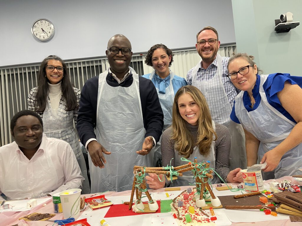 A smiling team proudly stands behind their creative chocolate bridge structure during The Chocolate Challenge team building event. Each team member is wearing aprons, and the table is filled with candy decorations and construction materials, showing their collaborative efforts.