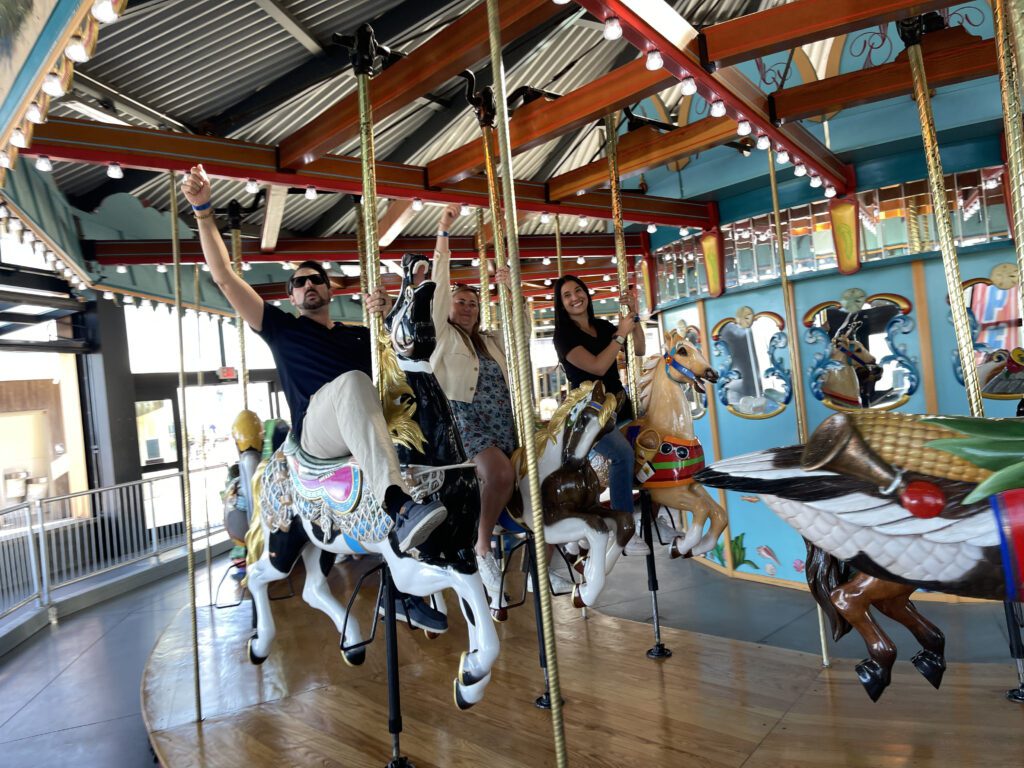 Participants enjoy a ride on a colorful carousel during a Custom Team Building event. They smile and wave as they ride the beautifully crafted horses, adding a playful touch to the experience.