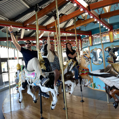 Participants enjoy a ride on a colorful carousel during a Custom Team Building event. They smile and wave as they ride the beautifully crafted horses, adding a playful touch to the experience.