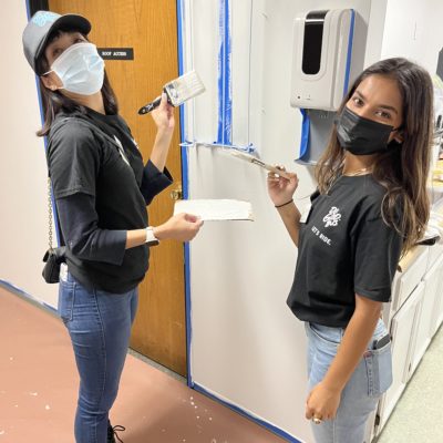 Two women are participating in a charitable team building event. They are painting an indoor wall together. Both are wearing black shirts and face masks. One woman on the left, wearing a black cap, holds a paintbrush and a paint tray, while the other woman on the right holds a paint roller. Blue painter's tape marks off the area they are painting. The setting appears to be an indoor facility, with a wooden door and a hand sanitizer dispenser mounted on the wall in the background.