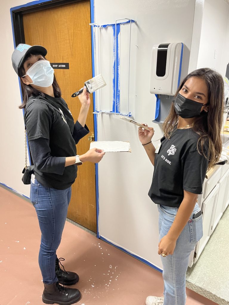 Two women are participating in a charitable team building event. They are painting an indoor wall together. Both are wearing black shirts and face masks. One woman on the left, wearing a black cap, holds a paintbrush and a paint tray, while the other woman on the right holds a paint roller. Blue painter's tape marks off the area they are painting. The setting appears to be an indoor facility, with a wooden door and a hand sanitizer dispenser mounted on the wall in the background.