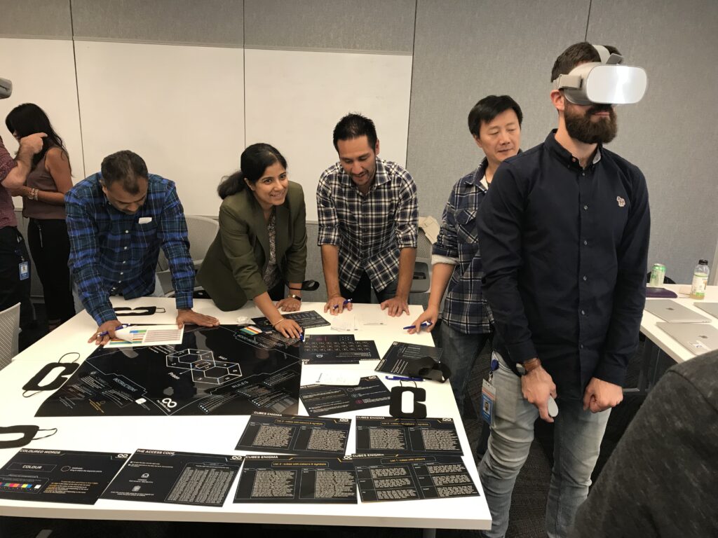 Team members collaborate at a table while one participant engages with a virtual reality headset during The Infinite Loop team building event.