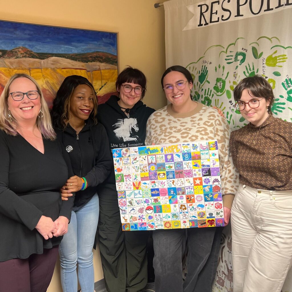 Group of participants smiling and holding a colorful mini-mural made of small painted tiles during the Make-A-Mini-Mural activity at a corporate charitable team building event called 'the Donation Station'.