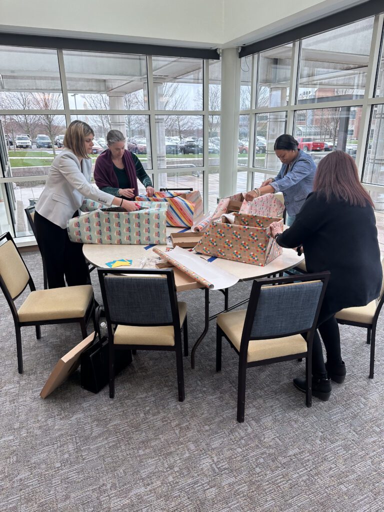 Four people are gathered around a table wrapping large boxes with colorful gift paper, creating a festive atmosphere as they prepare for birthday kits to donate as part of a team building event.