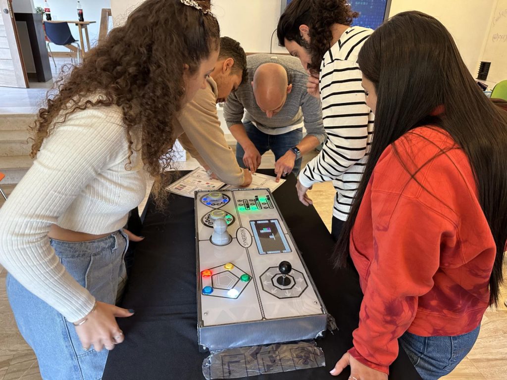 Team members gather around the Team Synergy gaming console, collaborating and strategizing during a hands-on team building activity.