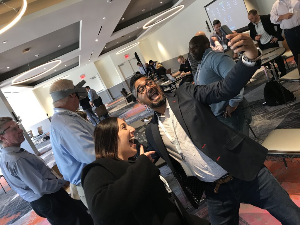 Two participants enthusiastically taking a selfie during The Infinite Loop team building event, while others engage in VR challenges in the background.