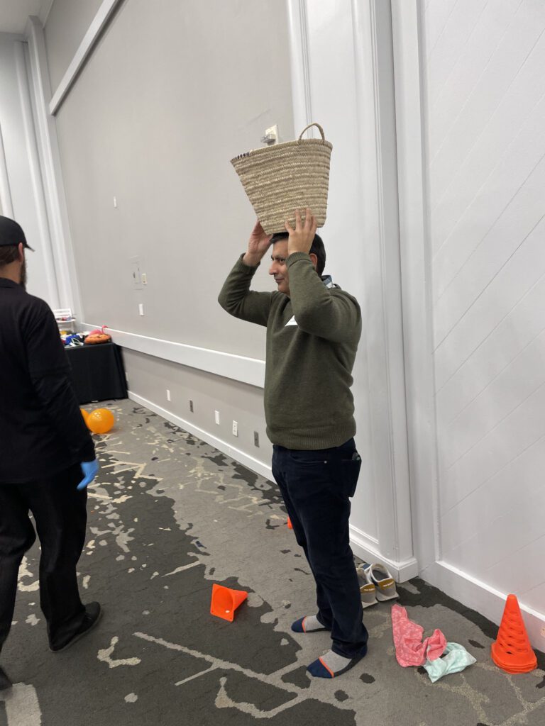 A man participates in a water walk challenge by balancing a basket on his head during a Clean Water Connection team building event. The activity mimics the experience of carrying water over long distances, which is a daily reality for many people in water-scarce regions. This hands-on challenge helps raise awareness about the global water crisis and the importance of providing clean water solutions.