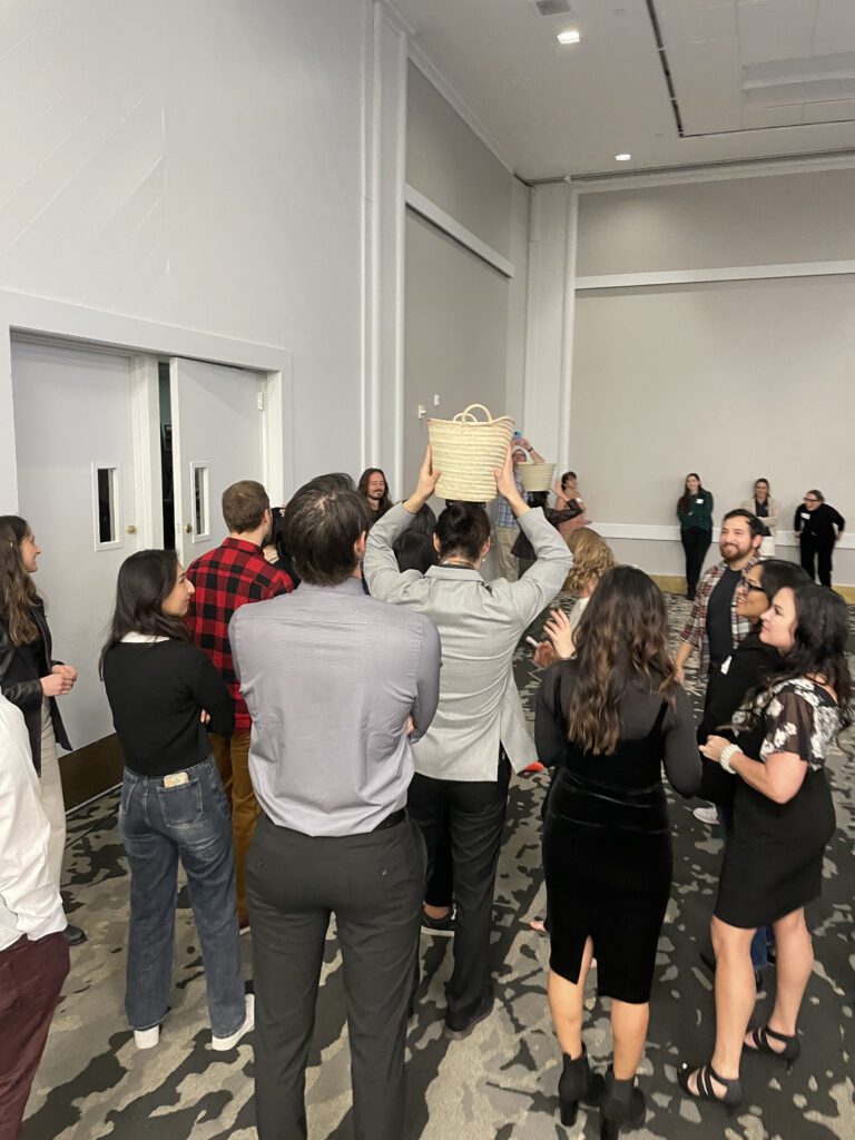 A group of participants observes as an individual balances a basket on their head during the water walk challenge at a Clean Water Connection event. This activity simulates the real-life experience of carrying water in underdeveloped regions, raising awareness about the global water crisis. The team building exercise promotes empathy and understanding while encouraging collaboration among participants.