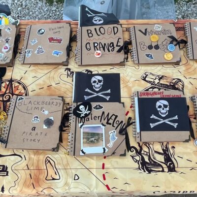 A table filled with pirate-themed notebooks decorated with stickers, skulls, and team names during a Custom Team Building event. The creative decorations reflect the playful pirate theme of the event.