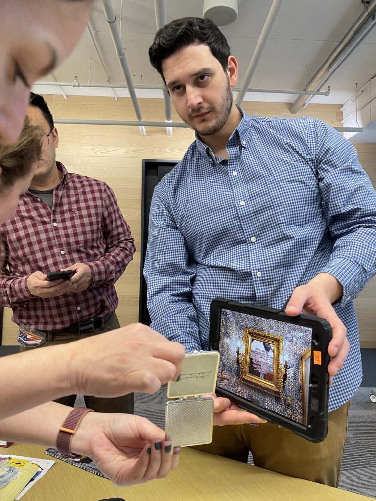 Team member holding a tablet showing a virtual crime scene as others examine a small evidence item during the Corporate CSI team building event.