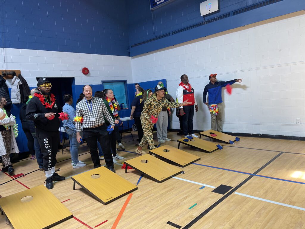 The image captures a group of participants engaging in a game of cornhole during an indoor team building event. The participants are standing behind cornhole boards, taking turns tossing bean bags toward the target.