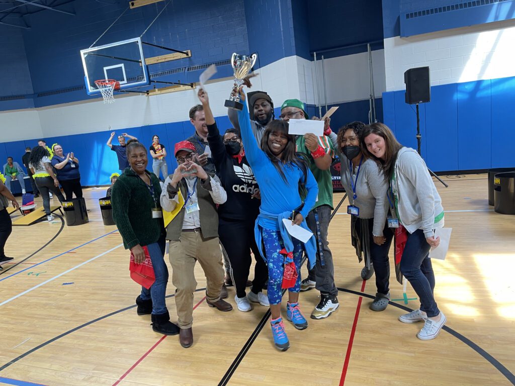 The image shows a group of participants celebrating a victory. The team is gathered in a gymnasium, smiling broadly and holding a trophy, signifying their success. Some members are holding certificates or awards, adding to the celebratory atmosphere. The group appears diverse, with people wearing casual attire, and some are wearing costumes or props, enhancing the fun, playful nature of the event. The joy and camaraderie evident in this image highlight the positive outcomes of team building activities, fostering a sense of unity and accomplishment among participants.