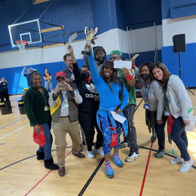 The image shows a group of participants celebrating a victory. The team is gathered in a gymnasium, smiling broadly and holding a trophy, signifying their success. Some members are holding certificates or awards, adding to the celebratory atmosphere. The group appears diverse, with people wearing casual attire, and some are wearing costumes or props, enhancing the fun, playful nature of the event. The joy and camaraderie evident in this image highlight the positive outcomes of team building activities, fostering a sense of unity and accomplishment among participants.