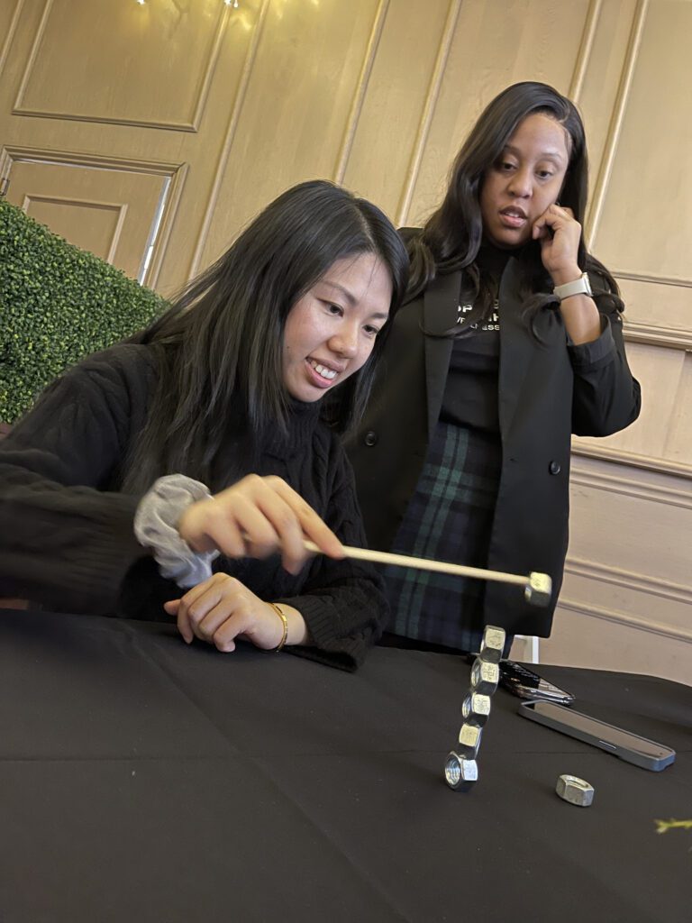 A participant skillfully balancing metal nuts in a vertical stack using a chopstick, while another participant watches with a look of concentration, during the In It to Win It team building event.