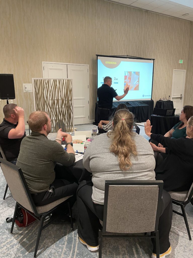 Participants attentively listen to a facilitator explaining striking techniques during a board-breaking team building session focused on overcoming obstacles and personal growth.