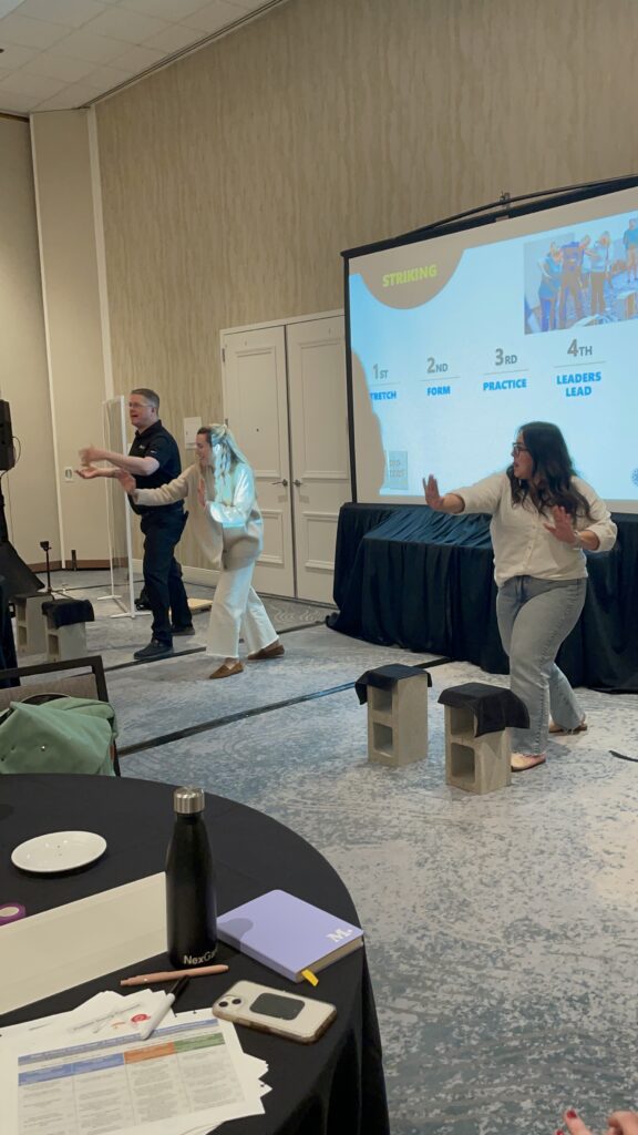 Two participants practice board-breaking techniques on stage during a team building event, guided by a facilitator.