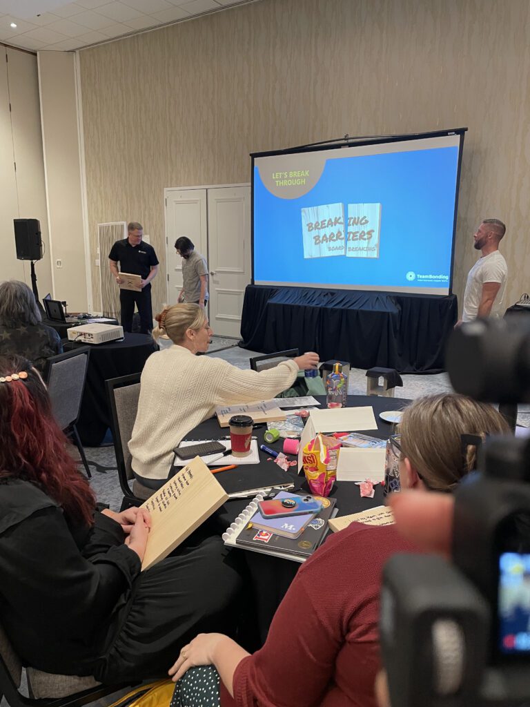 Participants sit at tables holding wooden boards while watching a facilitator prepare for the board-breaking demonstration during a team building event focused on breaking through barriers.