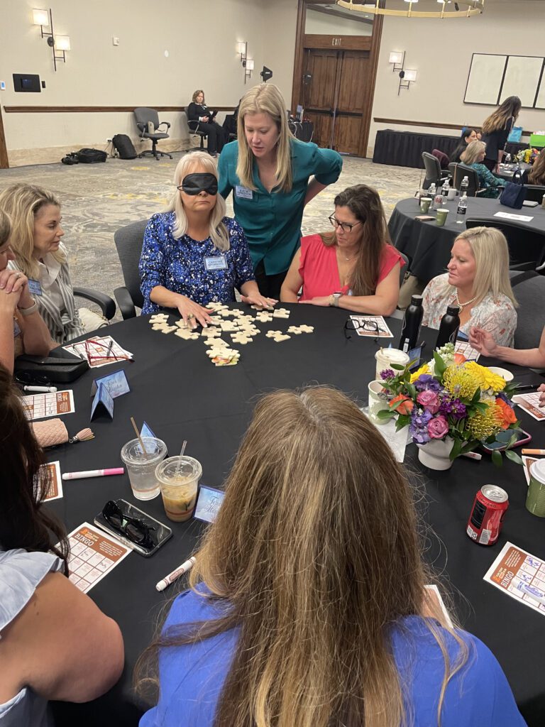 A group of women participating in a team building activity sitting around a round table. One woman is wearing a blindfold and feeling puzzle pieces in front of her. Other participants are observing and giving instructions to hep her complete the challenge.