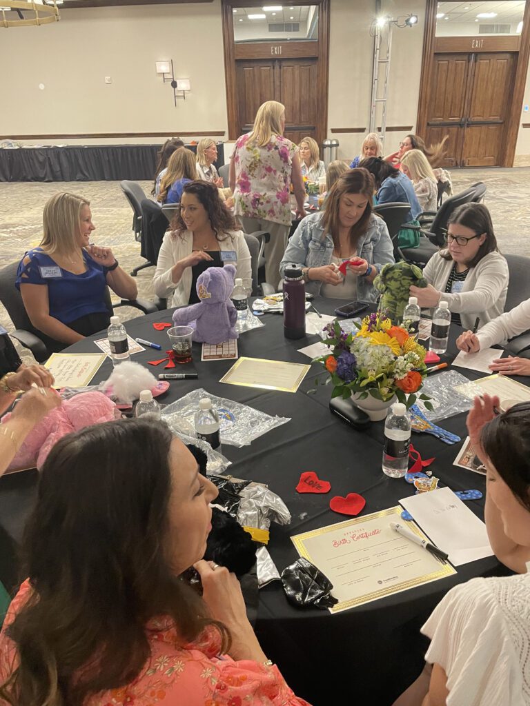 A group of women seated at a round table, engaged in a creative activity. Some are working on decorating or assembling colorful teddy bears, while others are chatting or focused on materials in front of them. The table is adorned with a vase of flowers, water bottles, and various teddy bear-making supplies, such as fabric hearts and stuffing. The room in the background shows more tables with participants working together in CSR team building program.