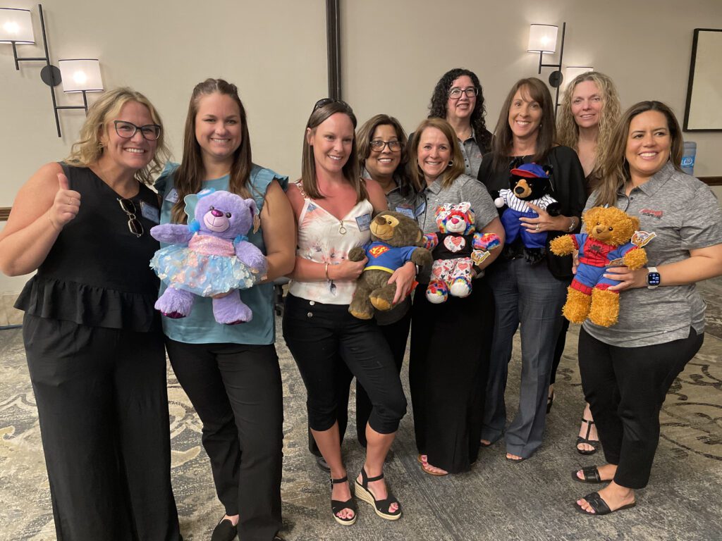 A group of eight smiling women posing together, holding customized teddy bears. Each teddy bear is dressed uniquely, with some wearing superhero costumes, a tutu, or colorful accessories. The women are standing in a conference room, showing off their creations proudly as part of a team building activity.