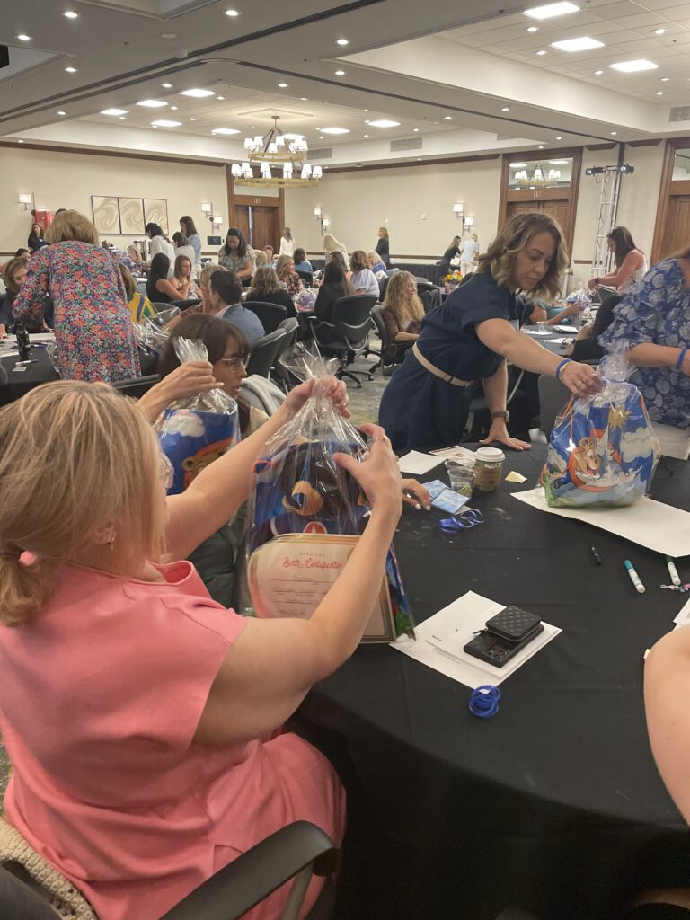 A group of women at a table are carefully assembling gift bags during a large event. The bags contain stuffed animals, certificates, and other items, for a charitable team building program. The room is filled with other participants engaged in similar activities.