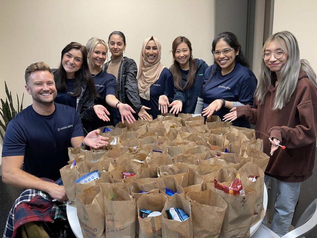 A group of eight smiling individuals stand around a table filled with brown paper bags, likely packed with food or care items. They are proudly showing off their work, extending their hands toward the bags. Everyone in the group appears happy and engaged in this Do Good Bus team building event.