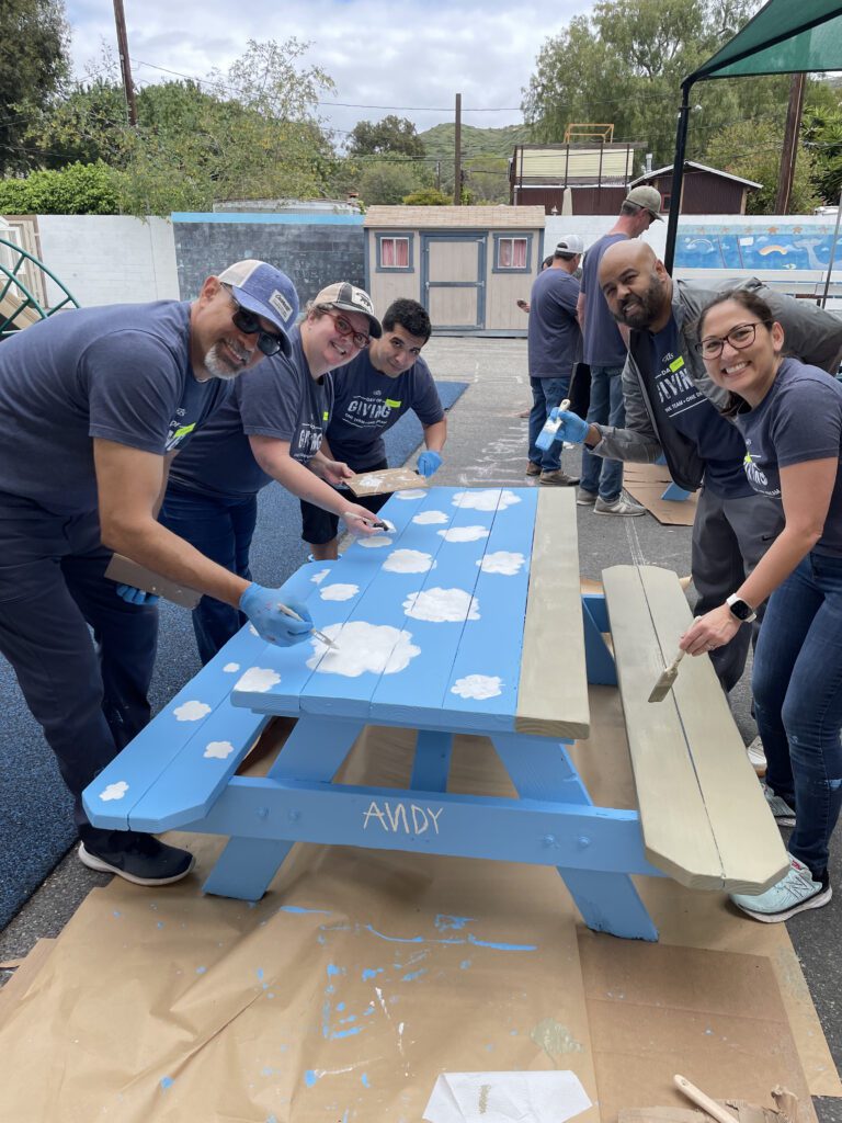 A group of five volunteers working together to paint an outdoor picnic table at a charitable team building event. The table is painted in bright blue with white clouds. All participants, wearing matching grey shirts and blue gloves, are smiling as they contribute to the project.