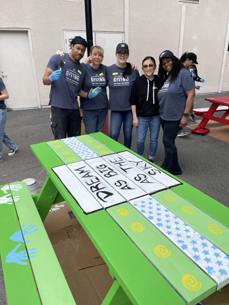 A group of five people standing together, smiling in front of a brightly painted green picnic table. The table is decorated with a motivational message, 