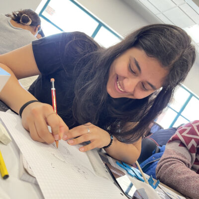 A young woman is smiling and fully engaged in drawing on graph paper. She appears focused and enjoying the creative process. She is holding a pencil and working on a sketch, with other materials spread out around her on the table.