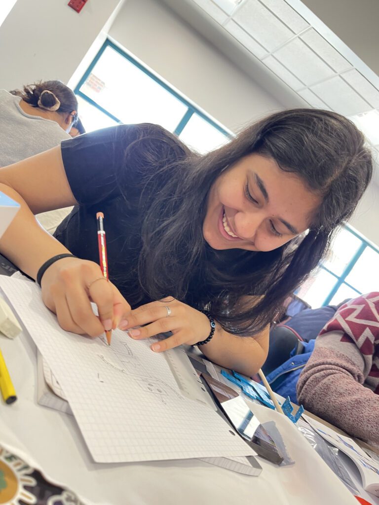 A young woman is smiling and fully engaged in drawing on graph paper. She appears focused and enjoying the creative process. She is holding a pencil and working on a sketch, with other materials spread out around her on the table.