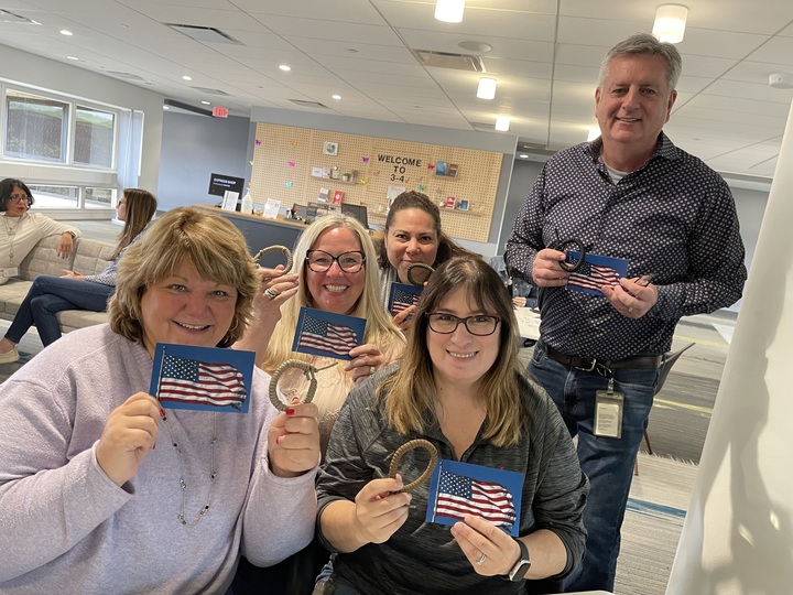 Group of participants smiling and holding paracord bracelets and cards with an American flag during the Paracord Bracelet Making activity at the corporate team building event 'Donation Station'.