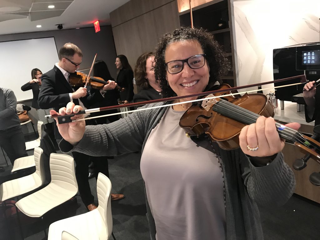 Smiling participant holds a violin and bow proudly during the Crescendo team building event, surrounded by teammates learning to play instruments.