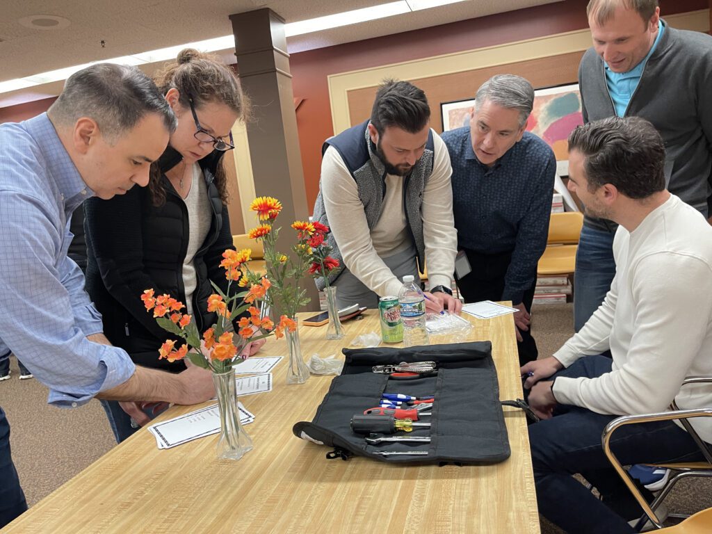 Group of participants gathered around a table during a Charity Bike Build event, reviewing instructions and using tools for bike assembly. A toolkit with screwdrivers and pliers is laid out on the table, and vases with colorful flowers add a decorative touch to the scene. The participants appear engaged in discussion while working collaboratively.