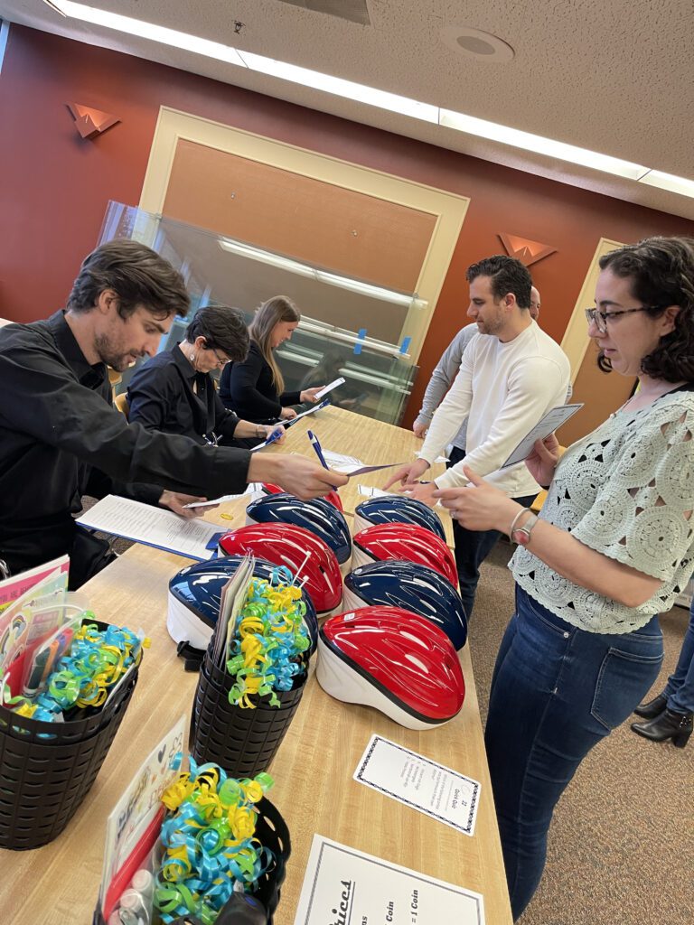 Participants in a team building event stand at a table with red and blue bicycle helmets, discussing instructions and checking documents. Gift baskets with colorful decorations are also displayed, highlighting the preparation phase of a Charity Bike Build event.