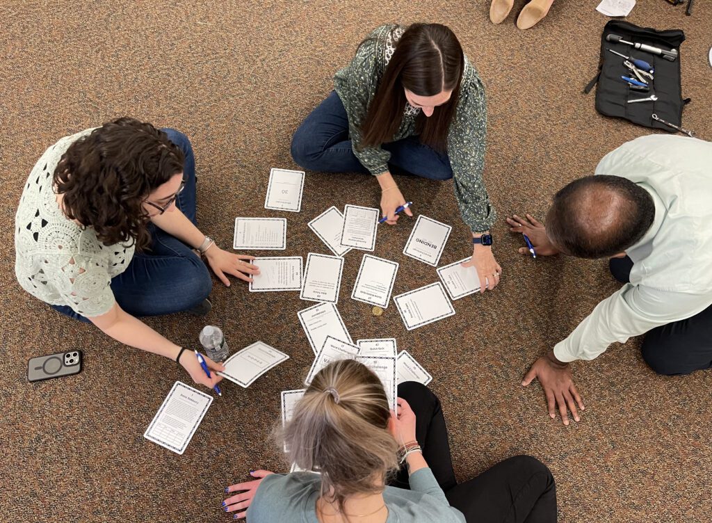 Four participants in the Charity Bike Build sit on the floor in a circle, collaborating as they fill out cards laid out in the center. The scene showcases teamwork and communication as they work together to complete this step in the bike-building process.