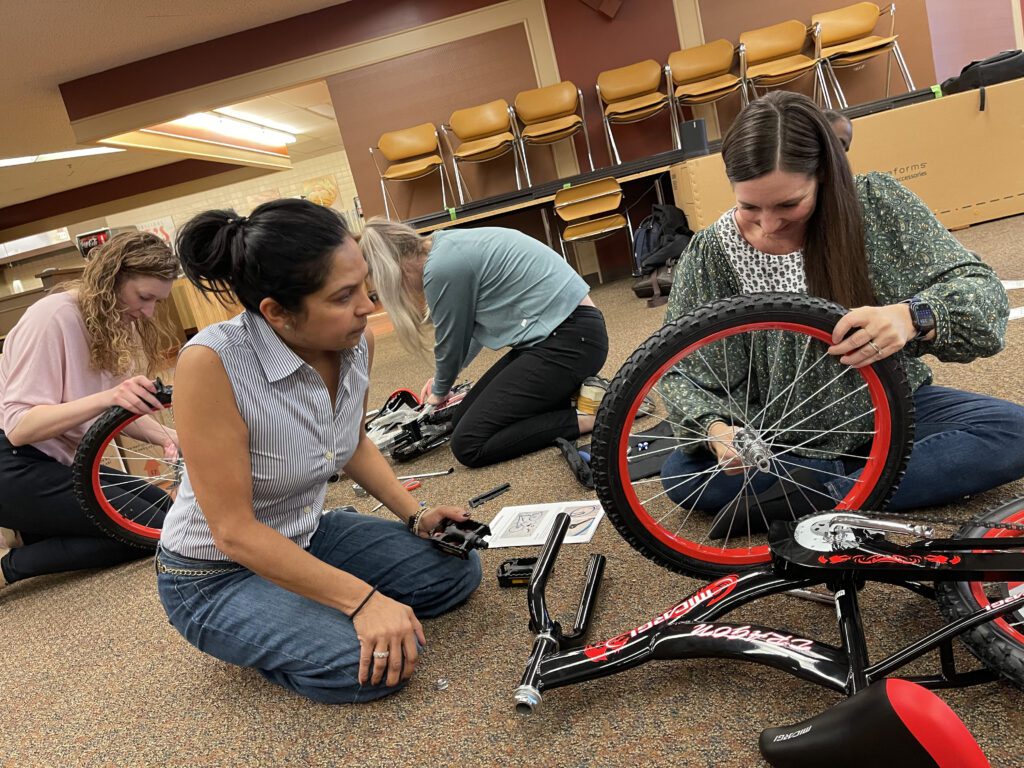 Four team members work together on the floor to assemble bikes during the Charity Bike Build event. They are focused on attaching wheels and following instructions, demonstrating collaboration and problem-solving as part of this hands-on, team building activity.